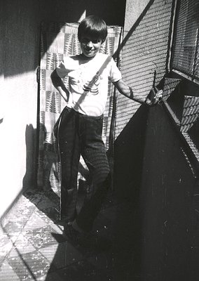 Young boy in 1960s-70s streetwear poses with acoustic guitar on urban staircase. Lighting suggests midday sun casting shadows...