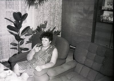 Vintage black-and-white portrait of a woman seated on a curved sofa in a mid-century interior, holding a cigarette. She wears...