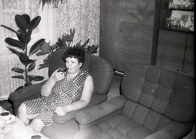 Mid-century indoor portrait of a woman in a polka-dot dress, seated on a tufted armchair beside a potted plant. Soft focus an...