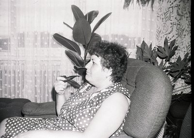 Mid-century indoor portrait of a woman in a polka-dot dress, seated in a reclined armchair beside a large leafy plant. The pa...