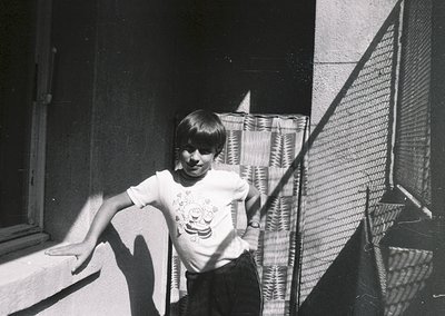 Young boy in 1970s-style short-sleeved shirt leans against a window ledge, framed by a mesh screen door. Indoor light casts s...