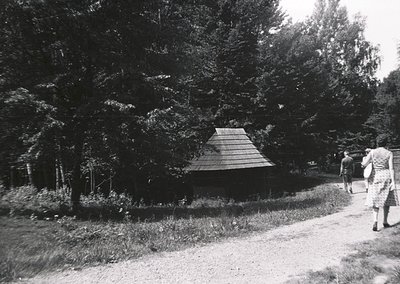 Mid-century wooden gazebo nestled in a forested area, surrounded by dense coniferous trees. Two figures in period attire (195...