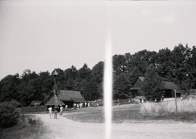 Black-and-white rural scene featuring a wooden pavilion with a conical roof, likely a folk architecture exhibit. Group of peo...