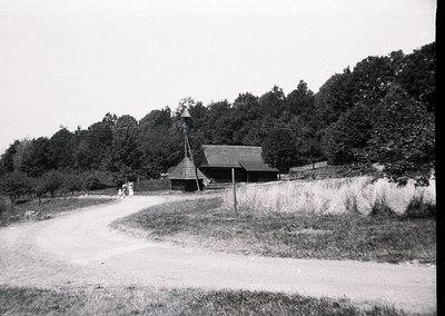 Rural countryside scene featuring a wooden barn with a steep gable roof and a small steeple, surrounded by dense forest. Two ...
