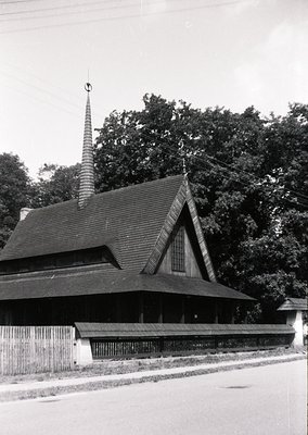 Wooden church with steep gable roof and tall, slender spire, likely Eastern European architecture. Rustic fence and mature tr...