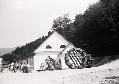 Historic waterwheel-powered mill in a rural alpine setting, likely 1940s-1950s. White triangular-roofed structure with arched...