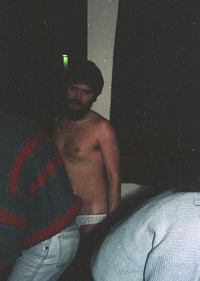 Vintage Polaroid-style photo of a shirtless man in low-light, holding a green fluorescent object. He wears light-colored pant...