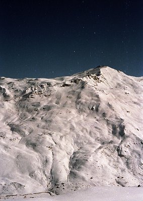 Snow-covered alpine peak under starry night sky, showcasing rugged terrain and dramatic lighting.