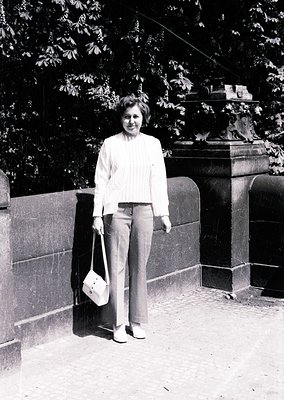 Mid-century woman in a light sweater and wide-leg trousers poses outdoors beside a stone wall and ornate lamp post. Classic 1...