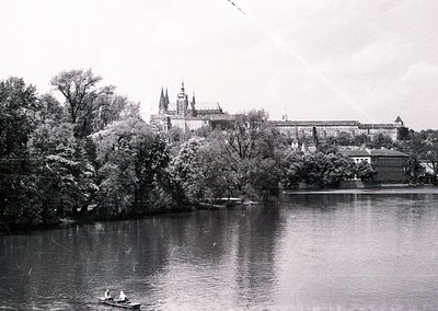 Historic black-and-white view of Prague Castle and the Vltava River, framed by autumn foliage. Gothic spires dominate the sky...