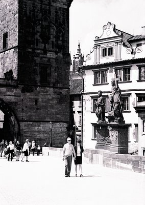 Historic Prague courtyard featuring Gothic stone archway and Baroque-style statue. Two figures in mid-20th-century attire (19...