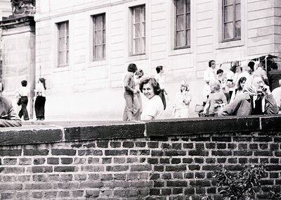 Black-and-white snapshot of a lively courtyard scene, likely 1960s–1970s. A group of people—mostly young—gather around a bric...