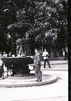 Classic 1960s fountain scene with ornate multi-tiered basin and sculpted figures. Central man in suit poses near water spout,...