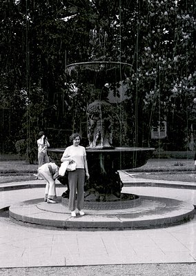 Classic 1970s-style fountain with sculpted figures and cascading water in a park setting. Two women pose near the center, one...