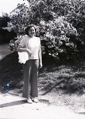 Mid-century woman in a striped sweater and wide-leg trousers stands outdoors, holding a rolled document or fabric. Lush green...