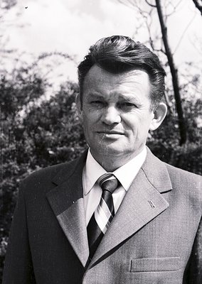 Portrait of a man in a sharp 1960s-70s suit, tie, and slicked-back hairstyle against an out-of-focus forest backdrop. Formal ...