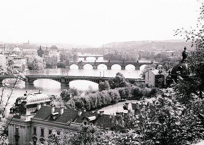 Black-and-white view of Prague’s Charles Bridge spanning the Vltava River, flanked by historic Baroque architecture. The brid...