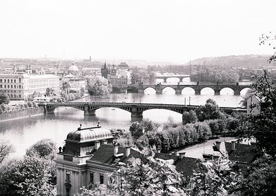 Vintage black-and-white view of Prague’s Charles Bridge spanning the Vltava River, flanked by historic Baroque and Gothic arc...