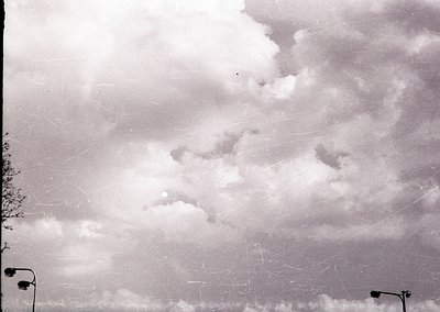 Vintage black-and-white aerial view of dense cloud cover with faint contrails crisscrossing sky. Partial urban landscape visi...