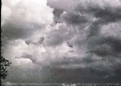 Vintage sepia-toned photograph of stormy skies with dramatic, billowing clouds and visible scratches/tears. Partial tree silh...