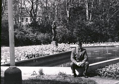 Mid-20th century man in formal attire sits by a reflective water feature in a landscaped park. Classical statue and manicured...