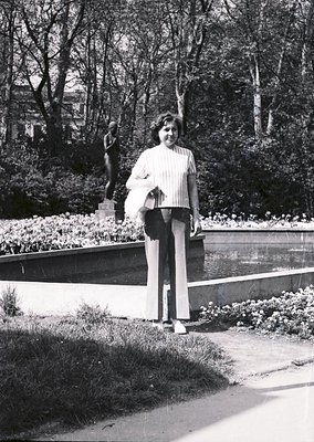 Black-and-white portrait of a woman in a 1970s park setting, holding a white bag. She wears a striped blouse, wide-leg trouse...