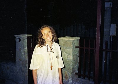 Young woman in a white shirt with a lanyard featuring a cross pendant, standing indoors near stone columns with intricate car...