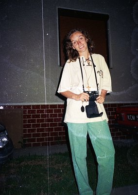 Vintage candid of a person holding a classic film camera, likely a Leica or similar, in front of a brick wall with graffiti. ...