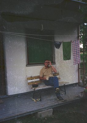 Man in casual 1970s attire rests on a wooden bench outside a weathered concrete building. Green window, green towel, and chec...