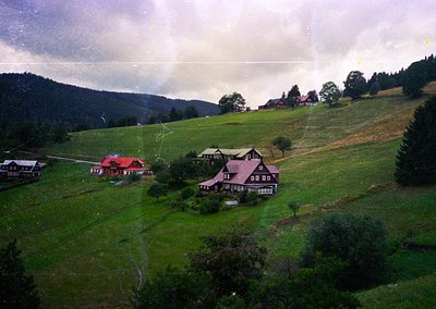 Aerial view of a rural Alpine village with clustered wooden chalets on rolling green hills. Central building features a red-t...