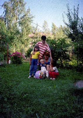 Family portrait in a lush garden setting, likely 1970s–1980s. Five individuals pose with a small dog and a gift-wrapped packa...