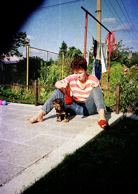 A person in a striped long-sleeve shirt and denim shorts sits on a concrete ledge, petting a small dog. Laundry hangs on a re...