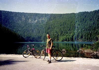 Retro-style photo of a man posing with two bicycles beside a serene alpine lake, framed by dense coniferous forests and snow-...