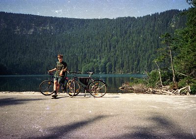 Vintage-style photo of a person posing with a classic mountain bike beside a serene alpine lake, surrounded by dense conifero...
