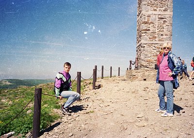 Two individuals pose atop a stone observation tower with panoramic valley views. The boy wears a purple jacket and backpack, ...