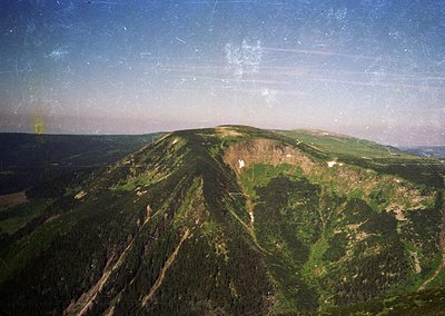 Aerial view of rugged alpine terrain with steep slopes and dense forestation. The image captures a mix of rocky outcrops and ...