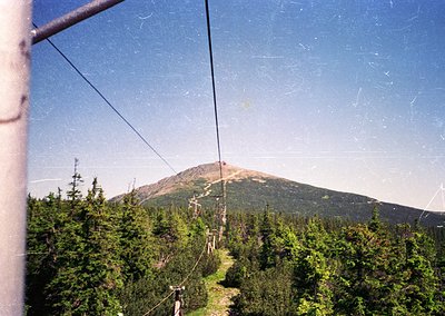 Vintage aerial tramway cable view of forested slopes leading to a snow-capped peak, framed by metal support structure. Dense ...