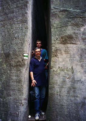 Two men pose in a narrow, weathered concrete tunnel. The man in front wears a blue polo shirt, jeans, and sneakers; the other...