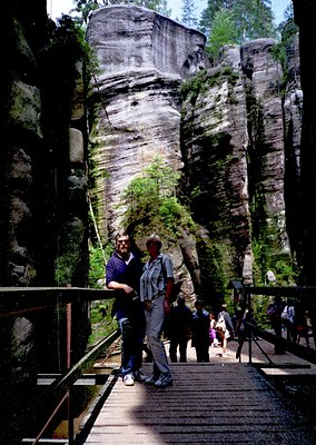 Two individuals pose on a wooden suspension bridge in a dramatic limestone canyon, framed by towering rock formations. The br...