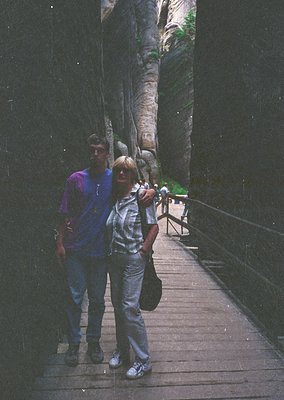 Two individuals pose on a wooden boardwalk flanked by rock walls, likely a canyon or gorge setting. The man wears a purple lo...