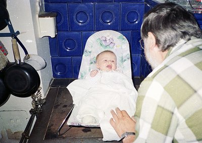 Vintage kitchen scene featuring a baby in a highchair with floral-patterned padding, swaddled in white. Adult male in checker...
