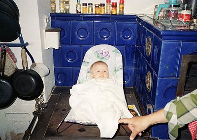 Vintage kitchen scene featuring a baby seated on a blue-tiled stove, wrapped in a floral-patterned cloth. Surrounding items i...