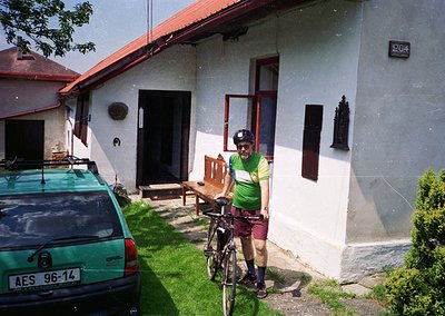 Cyclist in cycling gear poses beside a mid-20th-century residential building (, ). Light blue car (AES 96-14) parked adjacent...