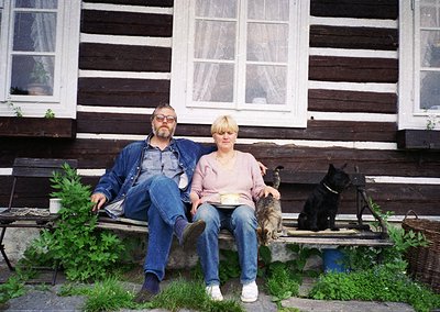 Couple posing on rustic wooden bench outside a weathered log cabin, flanked by two dogs. Denim attire and vintage camera sugg...