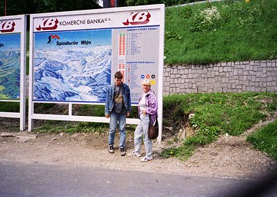 Two individuals pose beside a vintage Czech bank billboard advertising ski resort "Špindlerův Mlýn," featuring alpine imagery...