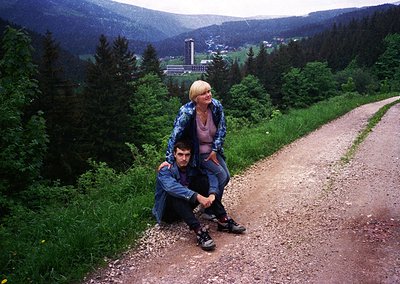 Couple posing on a gravel path in a lush alpine valley, surrounded by dense coniferous forests. Mid-20th century outdoor fash...