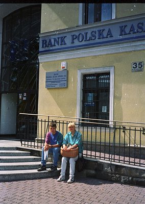 Two individuals sit on a wrought-iron railing outside a **Bank Polska Kasa** branch in a European city, likely **Warsaw, Pola...