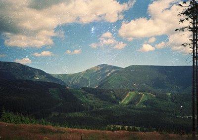 Vintage slide-style mountain landscape with dense coniferous forests, rolling terrain, and layered cloud formations. Evidence...
