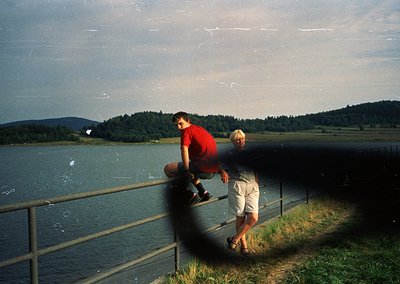 Two individuals pose on a metal railing beside a serene lake, framed by forested hills under a cloudy sky. The man in a red s...