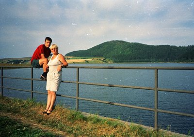 Couple posing on a lakeside railing, 1990s retro filter. Man in red polo, woman in white pantsuit. Green forested hill and ca...